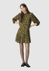 Sukienka w zielono-czarny wzór w panterkę z bufiastymi rękawami, falbaniastym dołem i wysokim kołnierzem. Modelka ma na sobie czarne baleriny i półprzezroczyste rajstopy.