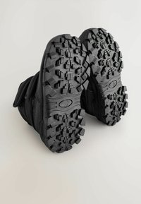 Sorte støvler med en tekstureret gummisål, der har et dybt mønster for bedre greb. Overdelen er lavet af slidstærkt stof og inkluderer velcro-remme.