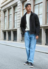 Jachetă bomber din suede negru, pulover tricotat crem, blugi drepți de culoare albastră deschisă, adidași negri cu talpă albă, fundal urban.