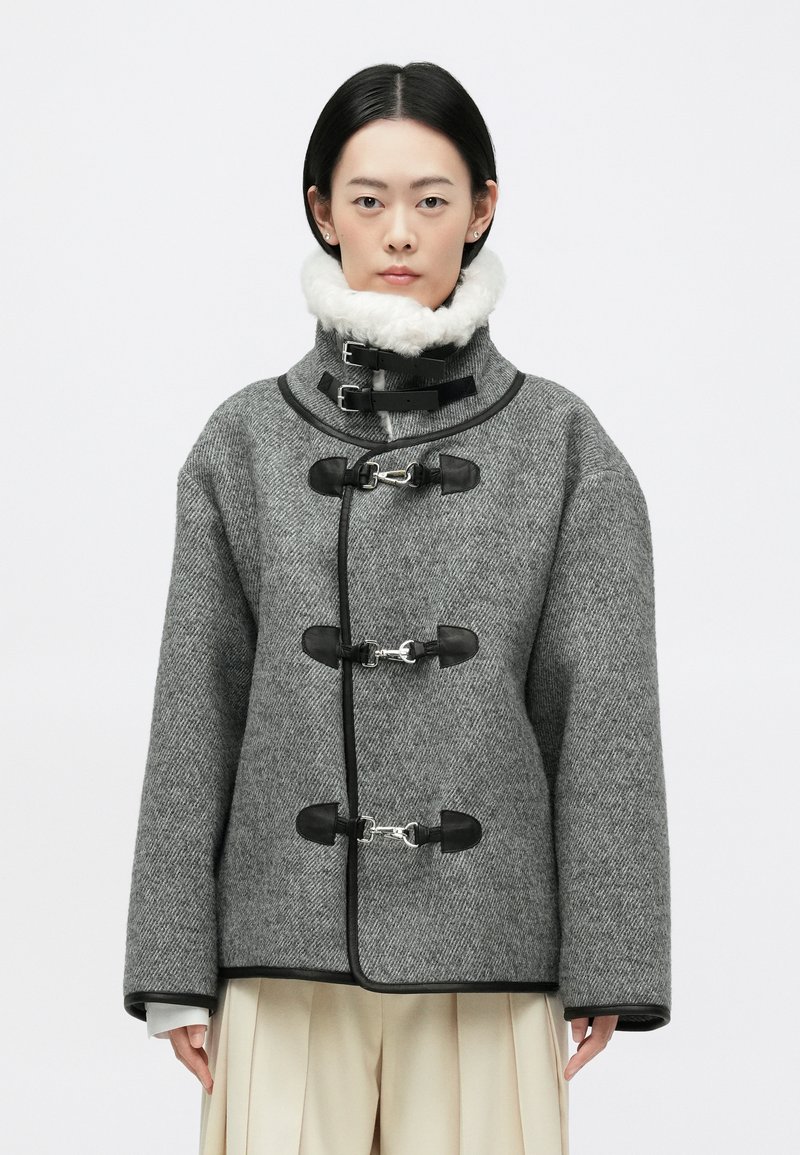 Sivi vuneni kaput s teksturiranim uzorkom riblje kosti, s crnim kožnim akcentima, velikim ovratnikom i zatvaranjem na dugmad. A-linija silueta s širokim rukavima.