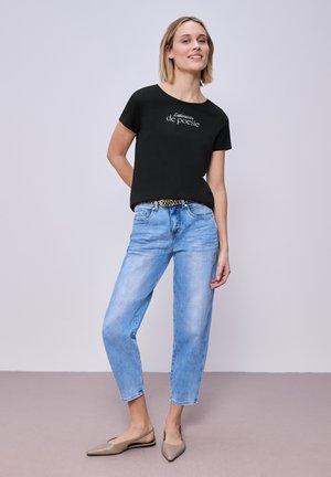 Frau lächelnd, trägt ein schwarzes "Laboratoire de poésie" T-Shirt, hellblaue Jeans mit Leopardenmuster-Gürtel und beigefarbene spitze Ballerinas, steht drinnen.