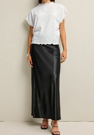 Vrouw draagt een gestructureerde witte top met korte mouwen en een lange, gladde zwarte rok met open schoenen, staand tegen een beige muur.