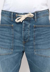Pantalones de mezclilla azul claro con cintura ajustable, que cuentan con dos grandes bolsillos frontales y costuras amarillas en contraste. Textura suave.