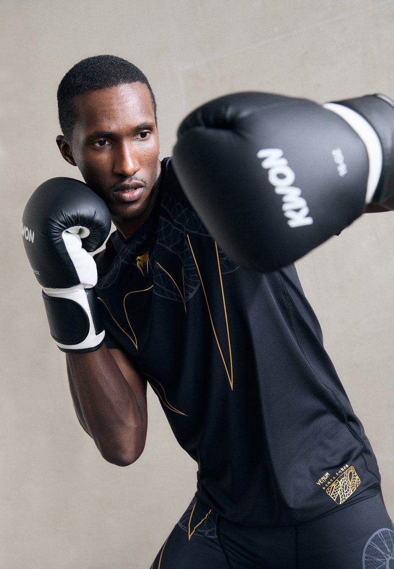 Boxeador en ropa deportiva negra y dorada, usando guantes de boxeo negros, levantando los puños en una postura defensiva y de golpeo contra un fondo neutral.