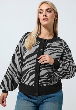 Donna con i capelli lunghi e biondi che indossa un cardigan con bottoni nero e grigio a stampa zebre e jeans blu, guarda di lato.