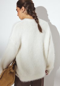 Pull en tricot blanc pelucheux avec une coupe ample, un col rond et des épaules tombantes. Texture douce avec un léger éclat. Porté avec un pantalon marron et un sac.