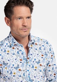 Chemise blanche avec des motifs floraux et de papillons bleus et orange, dotée d'un col évasé et d'une fermeture à boutons sur le devant. Texture lisse, tissu léger.