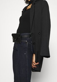 Svart kavaj med stora knappar över en svart figurnära topp, i kombination med mörkblå jeans. En liten svart midjeväska med lock sitter fast vid höften.