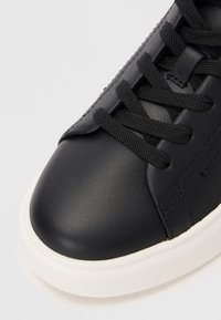 Adidași din piele neagră, cu vârful rotund, suprafață texturată și șireturi negre. Dispune de o talpă din cauciuc alb și detalii de cusătură subtile.