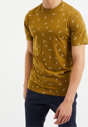 Man draagt een mosterdgele korte mouwt-shirt met kleine witte bladprint, gecombineerd met donkermarineblauwe broek, tegen een witte achtergrond.