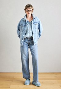 Lichtblauwe denimjacket met zakken, gedragen over een bijpassend shirt, gecombineerd met lichtblauwe wijde jeans en zachte blauwe schoenen.
