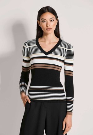 Vrouw met lang donker haar, gekleed in een zwart, wit en bruin gestreepte V-hals trui en zwarte broek, staand met één hand in haar broekzak.