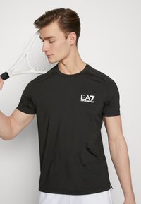 Camiseta atlética negra de mangas cortas con cuello redondo, que presenta un logo blanco de EA7 en el lado izquierdo del pecho y está hecha de una tela suave y ligera.