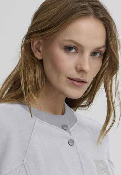 Light grey sweatshirt with a textured finish, featuring a round collar, button closures, and a subtle embroidered logo on the chest.