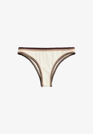 Femme aux longs cheveux foncés porte un bikini blanc avec une bordure marron, debout contre un mur rose texturé, touchant ses cheveux et regardant devant elle.
