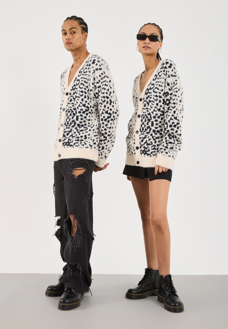 Cardigan tricoté en crème avec des taches de léopard noires, doté d'un col en V et de boutons. Les modèles portent des jeans noirs déchirés et des bottes noires.