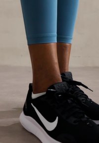 Pantofi sport negri Nike cu un swoosh alb, asortați cu colanți ajustați albaștri, evidențiind texturi fine și un design elegant.