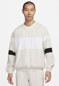 Pullover sportivo beige chiaro con strisce orizzontali bianche e nere, polsini elasticizzati e logo dorato Nike sul lato sinistro del petto.
