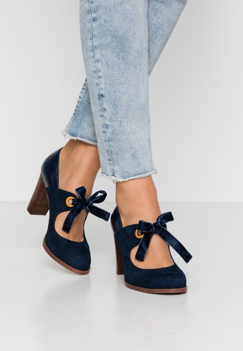 Chaussures à talons hauts bleu marine avec une finition en daim. Caractérisées par un design découpé, des nœuds en velours et un talon en bois. Portées avec un jean déchiré.