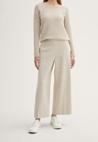 Beige strukturerad långärmad topp med en boxig passform, matchad med vida beige-byxor och vita sneakers, stående framför en neutral bakgrund.