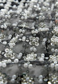 Schwarzer Spitzenstoff, verziert mit weißen Perlen und Pailletten, die florale Muster bilden. Die Textur ist komplex und geschichtet, was die Details betont.