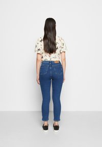 Calças skinny azul-escuras com uma cintura alta, apresentando dois bolsos traseiros e um patch de couro castanho na parte de trás. Usadas com uma blusa de manga curta estampada.