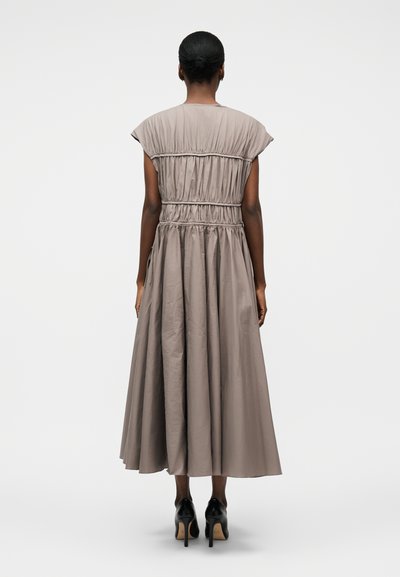 Žena stojaca chrbtom ku kamere, oblečená v taupe midi šatách s riasením, krátkymi rukávmi a čiernymi lodičkami na vysokom opätku.