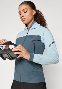 Šviesiai mėlyna ir tamsiai pilka striukė su gaubtu, užtrauktuko kišene, laikomomis rankogaliais ir logotipu; asmuo laiko slidinėjimo akinius. Strukturinė medžiaga.