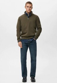 Sudadera verde oliva con cremallera y tejido texturizado, llevada sobre una camisa de mezclilla azul, combinada con unos vaqueros azul oscuro y zapatos negros.