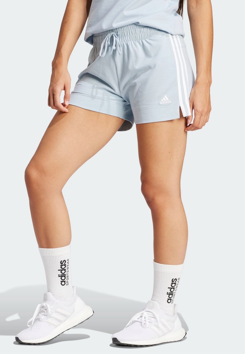 Frau in hellblauen Adidas-Shorts mit weißen Streifen, weißen Adidas-Crew-Socken und weißen Adidas-Laufschuhen.