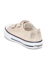 Zapatilla metálica dorada con diseño de corte bajo, suela de goma blanca y dos cintas de Velcro. Presenta una franja en contraste de color navy y el logo de la marca en el talón.