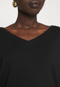 Czarny sweter w serek z prążkowanymi brzegami, wykonany z miękkiej dzianiny. Pokazuje zbliżenie na dekolt i gładką teksturę. Widoczne złote kolczyki w kształcie kół.