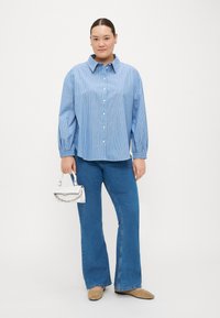 Camisa azul clara com riscas, com botões e colarinho, combinada com calças de ganga azuis largas. A modelo segura uma pequena mala branca com uma corrente.