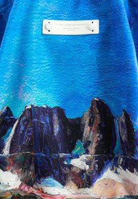 Prenda azul con un estampado texturizado de paisaje montañoso y una etiqueta blanca con dos broches circulares. El diseño resalta contrastes de color vibrantes.