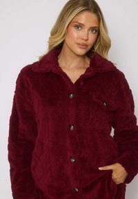 Bordeaux fleece jack met een gestructureerd ontwerp, knoopsluiting en een kraag. Beschikt over een zijzak en een comfortabele, zachte stof.