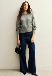 Grijze gebreide trui met geribde zoom, gecombineerd met donkerblauwe wijde jeans. Accessoires omvatten een kleine zwarte tas met een zilveren ketting.