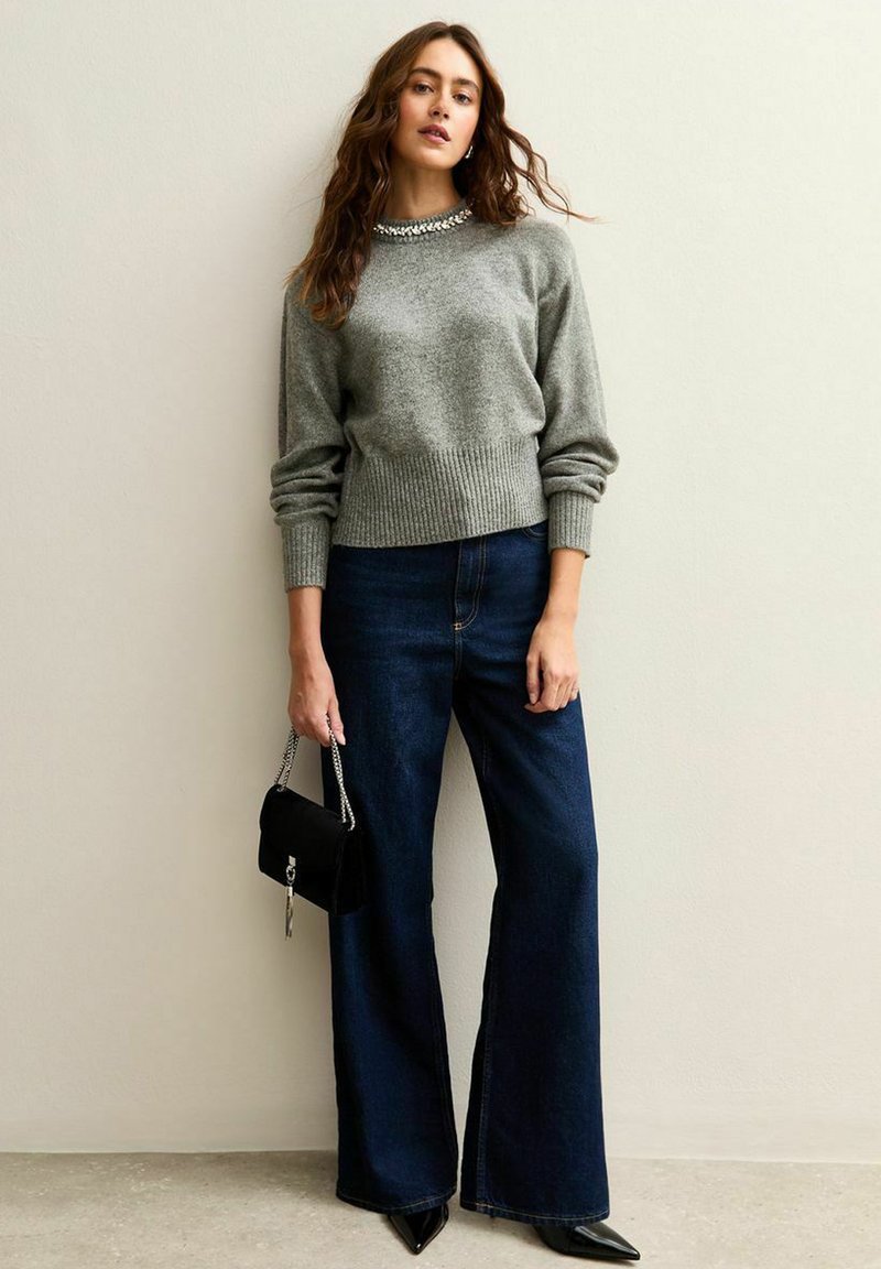 Grijze gebreide trui met geribde zoom, gecombineerd met donkerblauwe wijde jeans. Accessoires omvatten een kleine zwarte tas met een zilveren ketting.