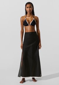 Sort bikini-top med hvid kant og sort gennemsigtig maxinederdel med høje slidser. Modellen står barfodet mod en neutral baggrund.