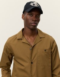 Barna gombos ing, mellzsebbel, fekete sapkával párosítva, amelyen fehér földgömb minta látható. Simább anyag és kényelmes szabás jellemzi.