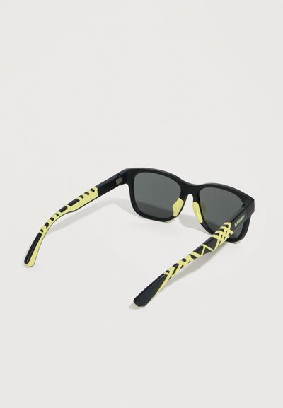 Ματ μαύρα γυαλιά ηλίου με σκοτεινούς φακούς, διακοσμημένα με νεον κίτρινα γεωμετρικά σχέδια στα πλαϊνά και νεον κίτρινες λεπτομέρειες στη γέφυρα της μύτης.