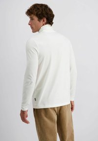 Man met krullend haar, gekleed in een witte lange mouwen turtleneck en lichtbruine corduroy broek, die naar links kijkt op een effen achtergrond.