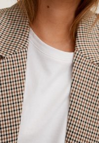 Hnědo-bílý kostkovaný blazer s klopovým límcem přes bílý tričko s kulatým výstřihem; viditelná textilie s přizpůsobeným střihem.
