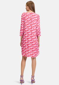 Rosa Kleid mit geometrischem Muster in verschiedenen Rosa- und Weißtönen. Es hat Dreiviertelärmel und eine lockere Passform. Getragen mit lila Absätzen.