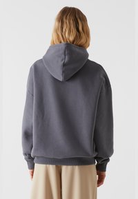 Mørkegrå hættetrøje med en stor hætte, drop shoulders og ribbede manchetter. Blød tekstur med en afslappet pasform, båret over lyse beige bukser.