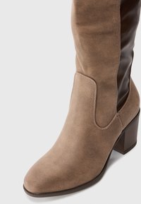 Taupe velúr bokacsizma tömbsarkúval és sötét rugalmas oldalpanellel egy sima fehér háttér előtt.