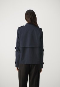Marineblauwe trenchcoat met een dubbelgestikte ontwerp, schouderepauletten en een gelaagde achterkant. Gladde textuur, lange mouwen en een getailleerde pasvorm.