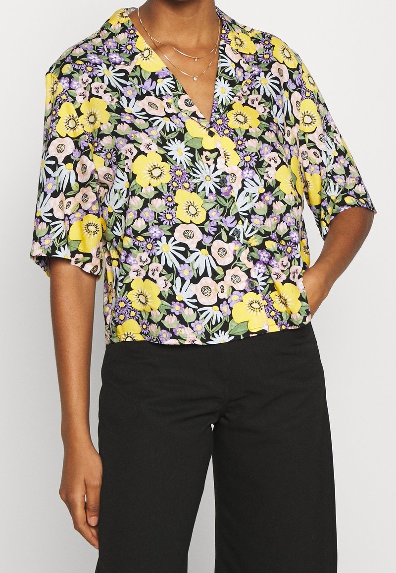 Blouse à manches courtes à motifs floraux mettant en avant un fond noir, avec des fleurs jaunes, roses et violettes éclatantes, dotée d'une coupe décontractée et d'un décolleté en V.