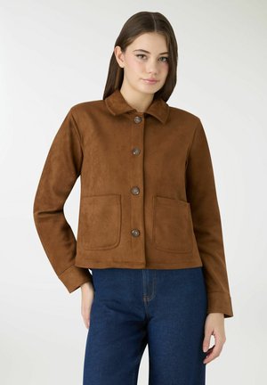 Giovane donna che indossa una giacca marrone con bottoni e grandi tasche frontali, con jeans blu, in piedi davanti a uno sfondo bianco uniforme.