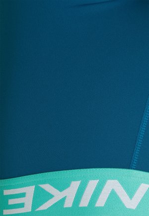 Niebieska powierzchnia materiału z turkusową elastyczną taśmą, na której znajduje się duży biały częściowy napis "NIKE" oraz widoczne szwy po prawej stronie.