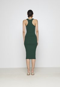 Abito midi a coste verde scuro con silhouette aderente, design a schiena scoperta e lunghezza fino al polpaccio, indossato con tacchi di colore chiaro.